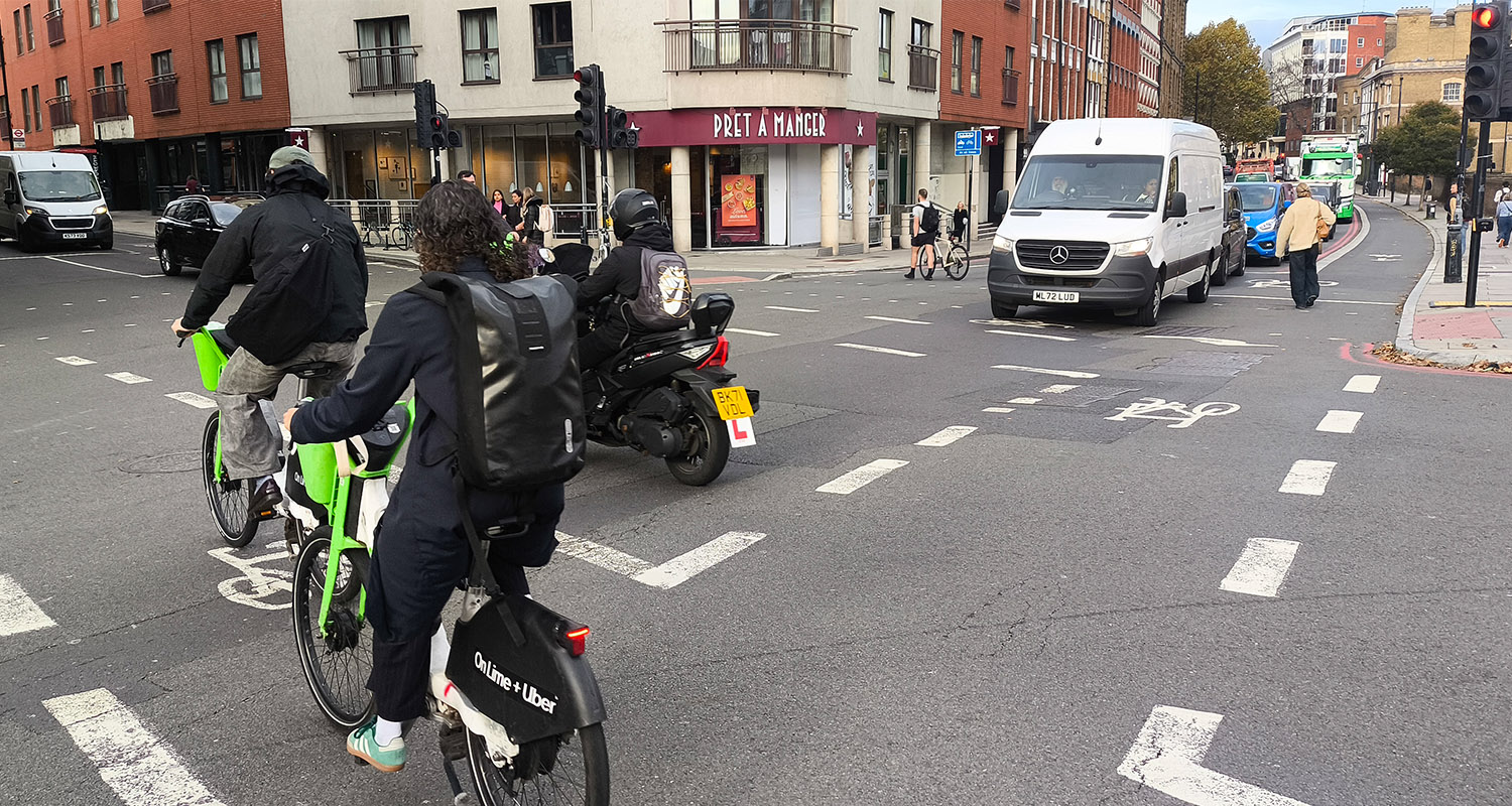 A junction in farringdon with painted cycle lanes across it but no protection. Two lime bikes and a motorbike are crossing while a white van is stopped in the middle of a pedestrian crossing at a red light.