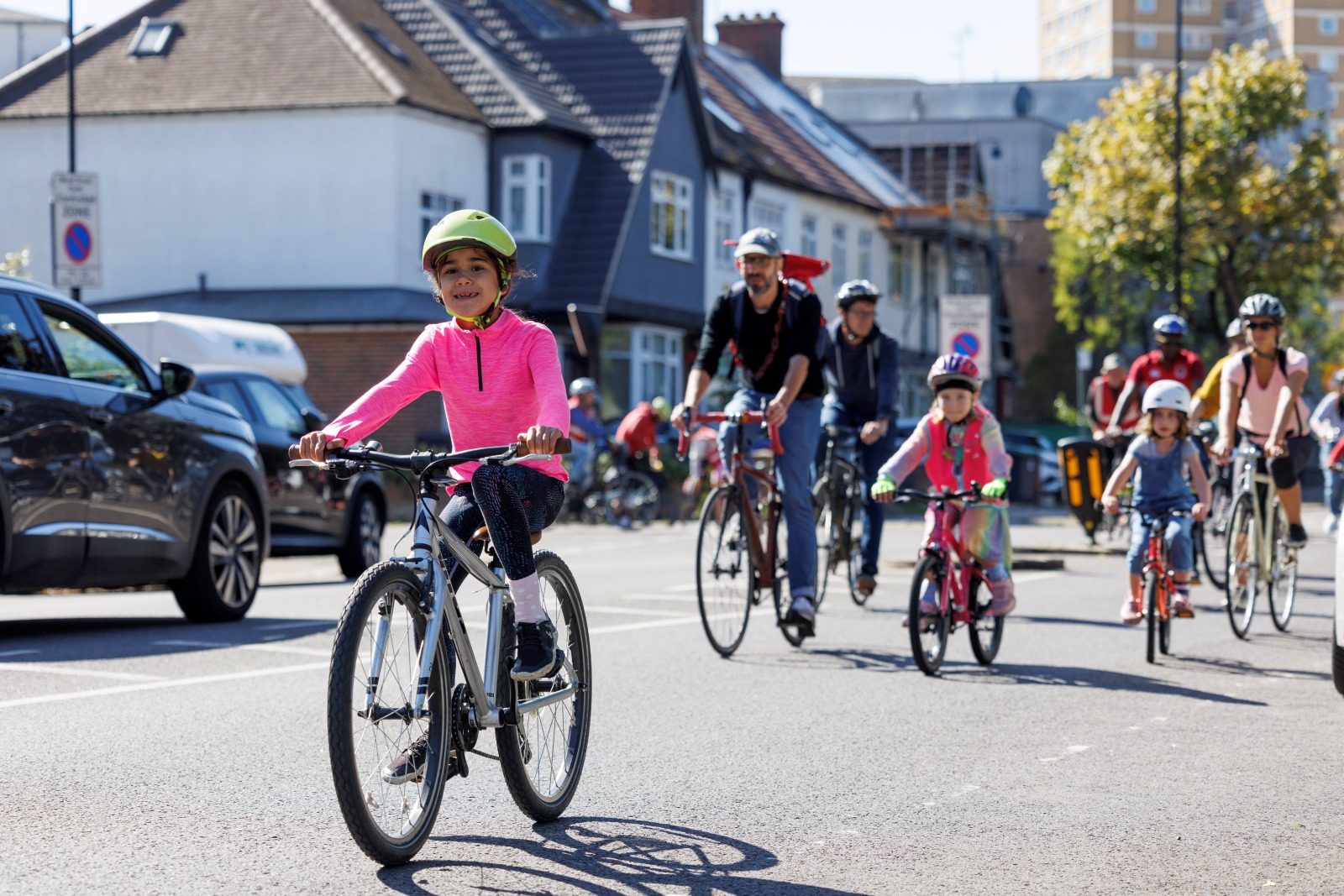A child cycling in a bright pink shirt and yellow helmet on a quiet street with other children and adults behind her.